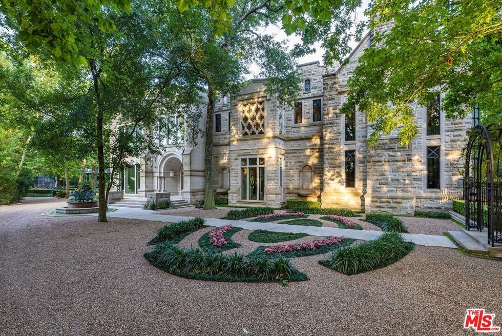 A wide, two-story stone house behind a gravel driveway and a tree.