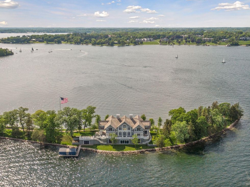 A three-story home on a narrow private island in the middle of a lake lined with trees.
